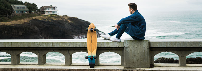 Guy sitting on bridge above ocean with Retrospec Zed Longboard leaning next to him