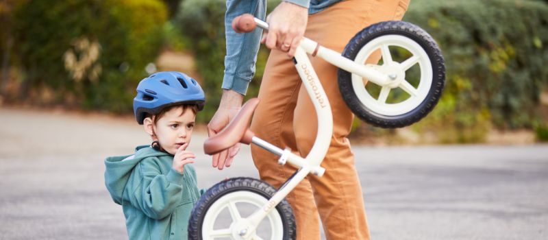 Person handing balance bike to a toddler wearing a helmet