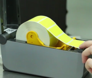 Photo of an open label printer with a hand grabbing yellow labels on a roll